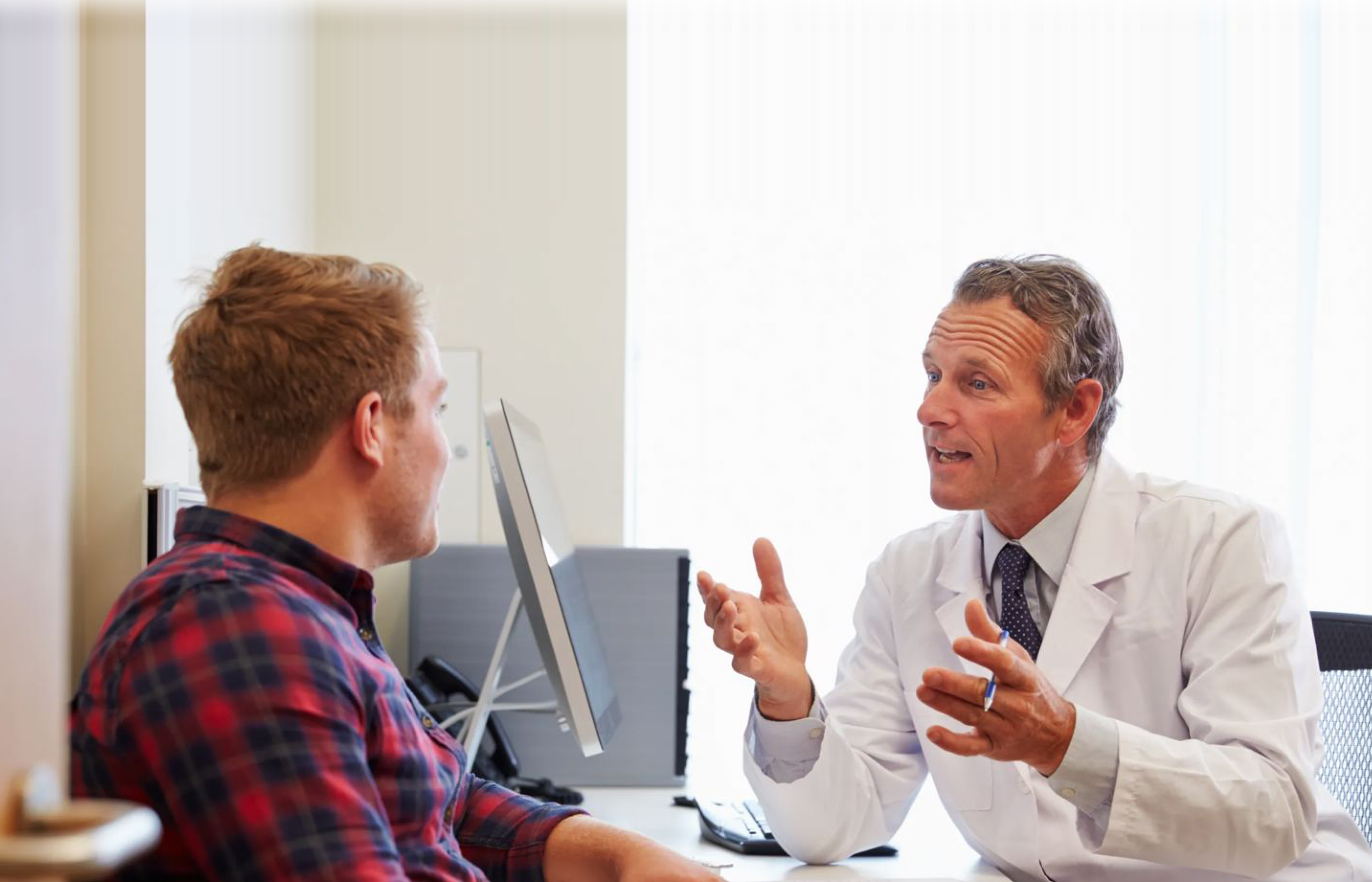 A doctor sitting next to a computer speaks with a male patient in an office.